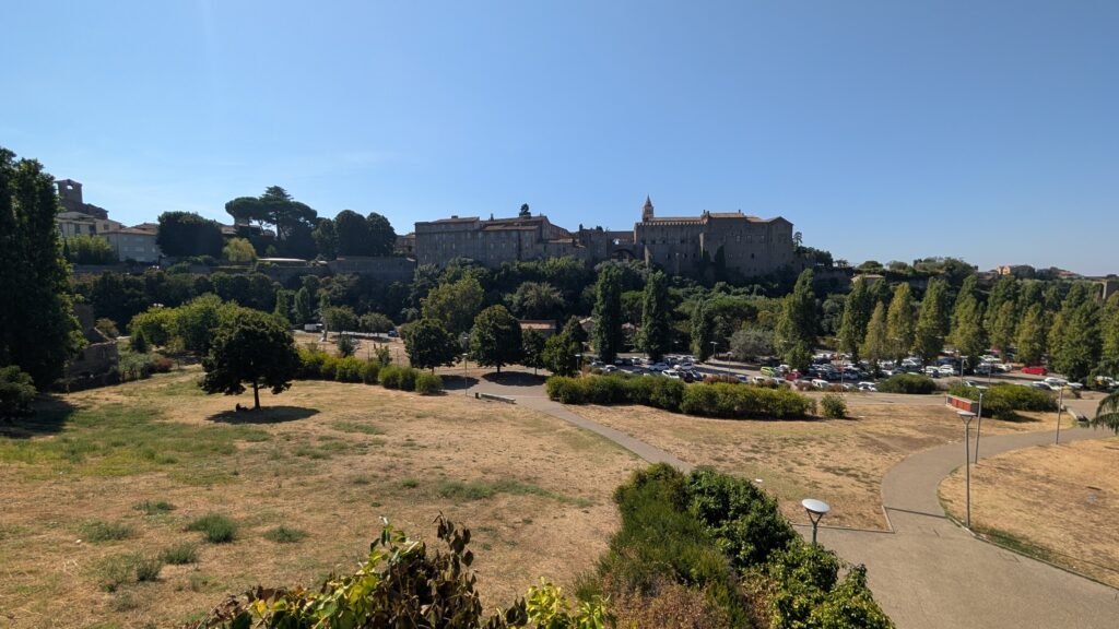 Valle Faul a Viterbo ad agosto: prato secco in primo piano, il parcheggio e il Palazzo dei Papi sullo sfondo