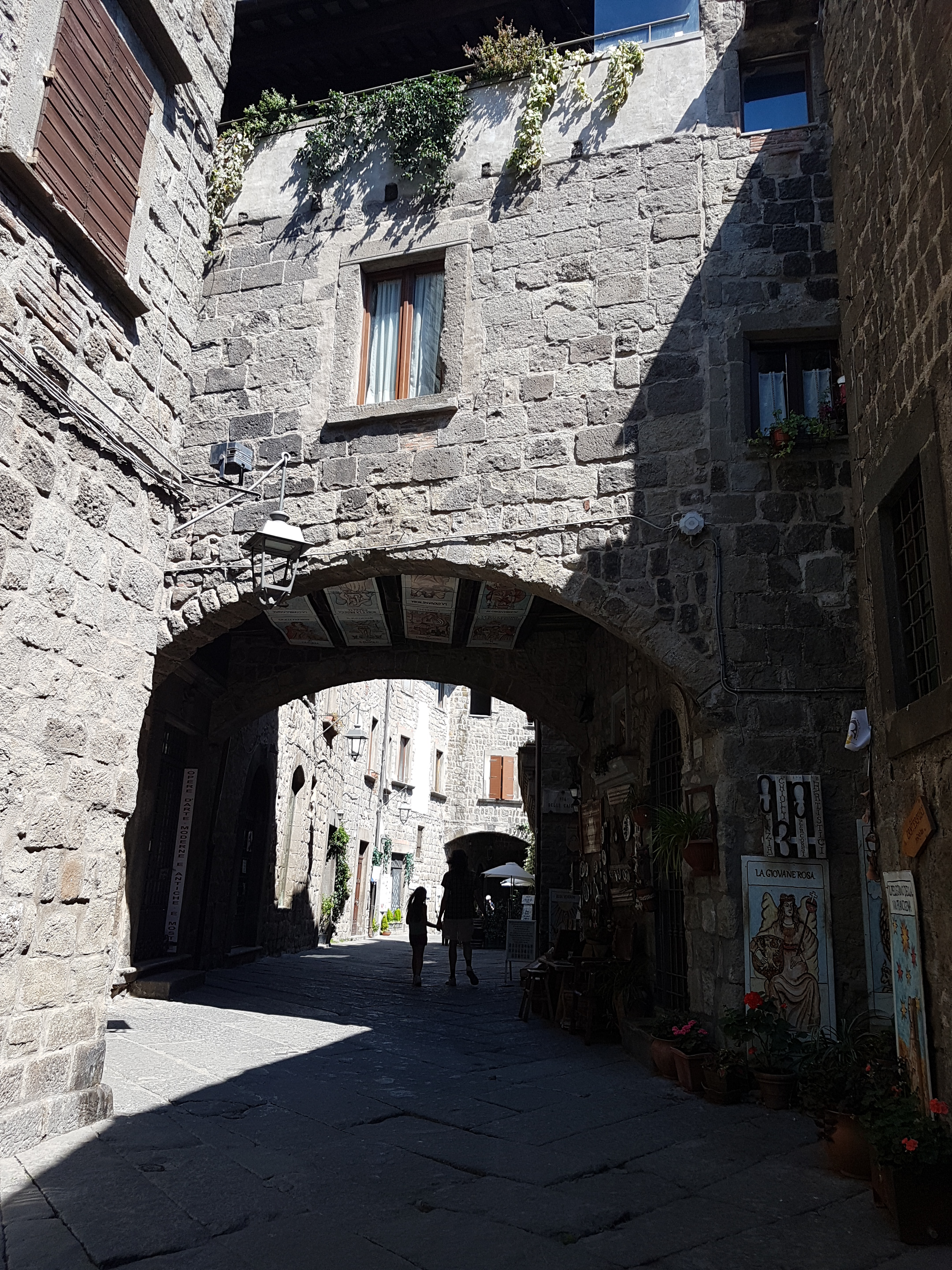 Un arco in peperino su una via lastricata del quartiere San Pellegrino, a Viterbo, con una bottega e due persone che vi passano sotto