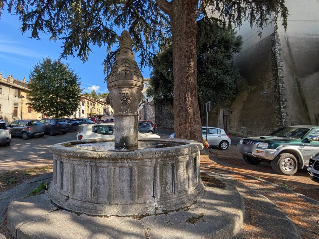 La fontana dell’Olmo, una colonna a fuso con musi di leone sopra una vasca circolare in pietra, sotto gli alberi di piazza Piccolomini
