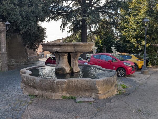 Fontana a coppa sorretta da pilastri sopra una vasca lobata, in piazza Piccolomini a San Martino al Cimino, con auto parcheggiate dietro