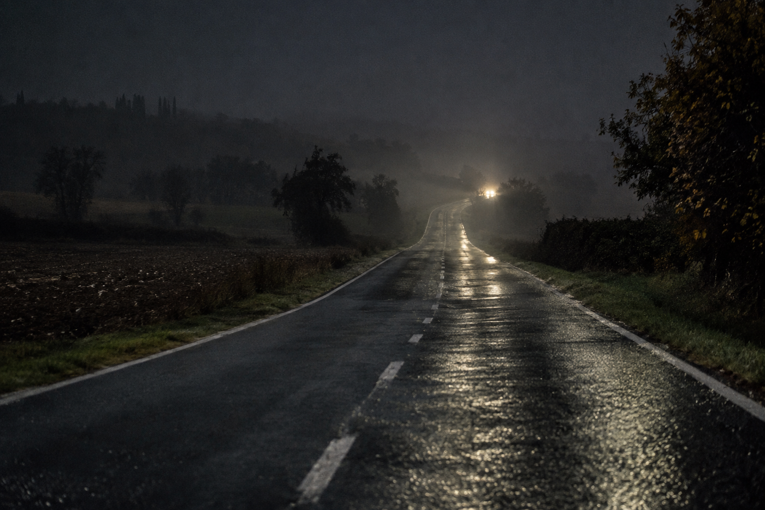 Strada di campagna deserta di notte, con i fari di un'auto in lontananza tra la nebbia