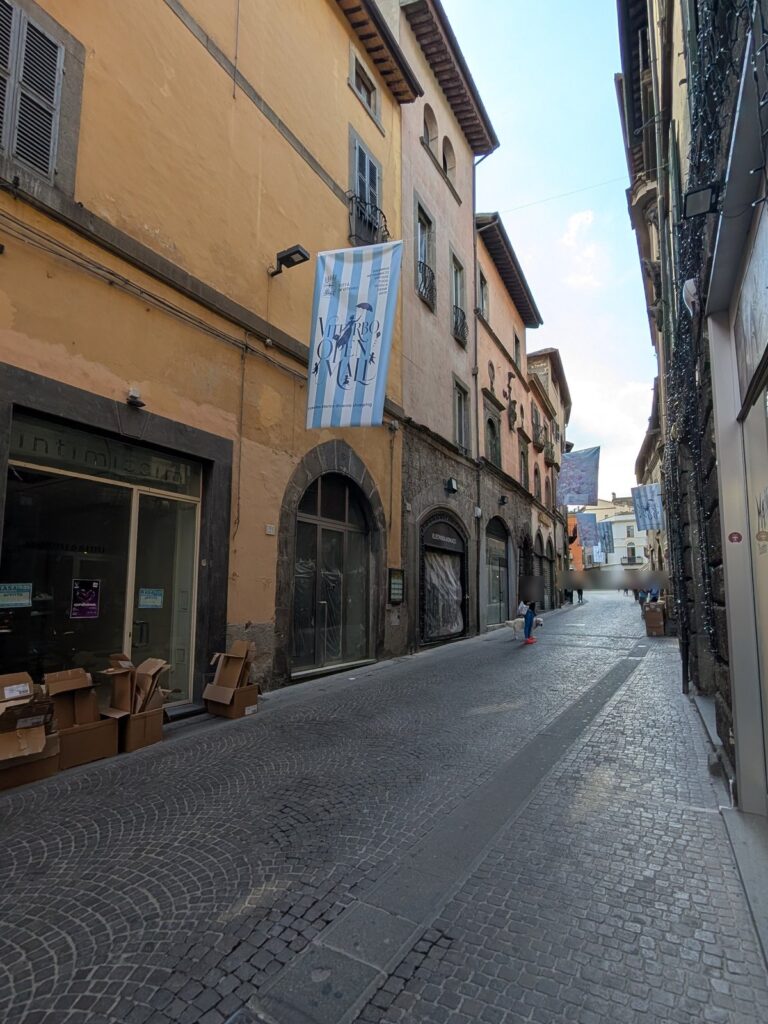 Una via del centro storico di Viterbo con uno stendardo di Viterbo Open Mall e vetrine lungo il percorso