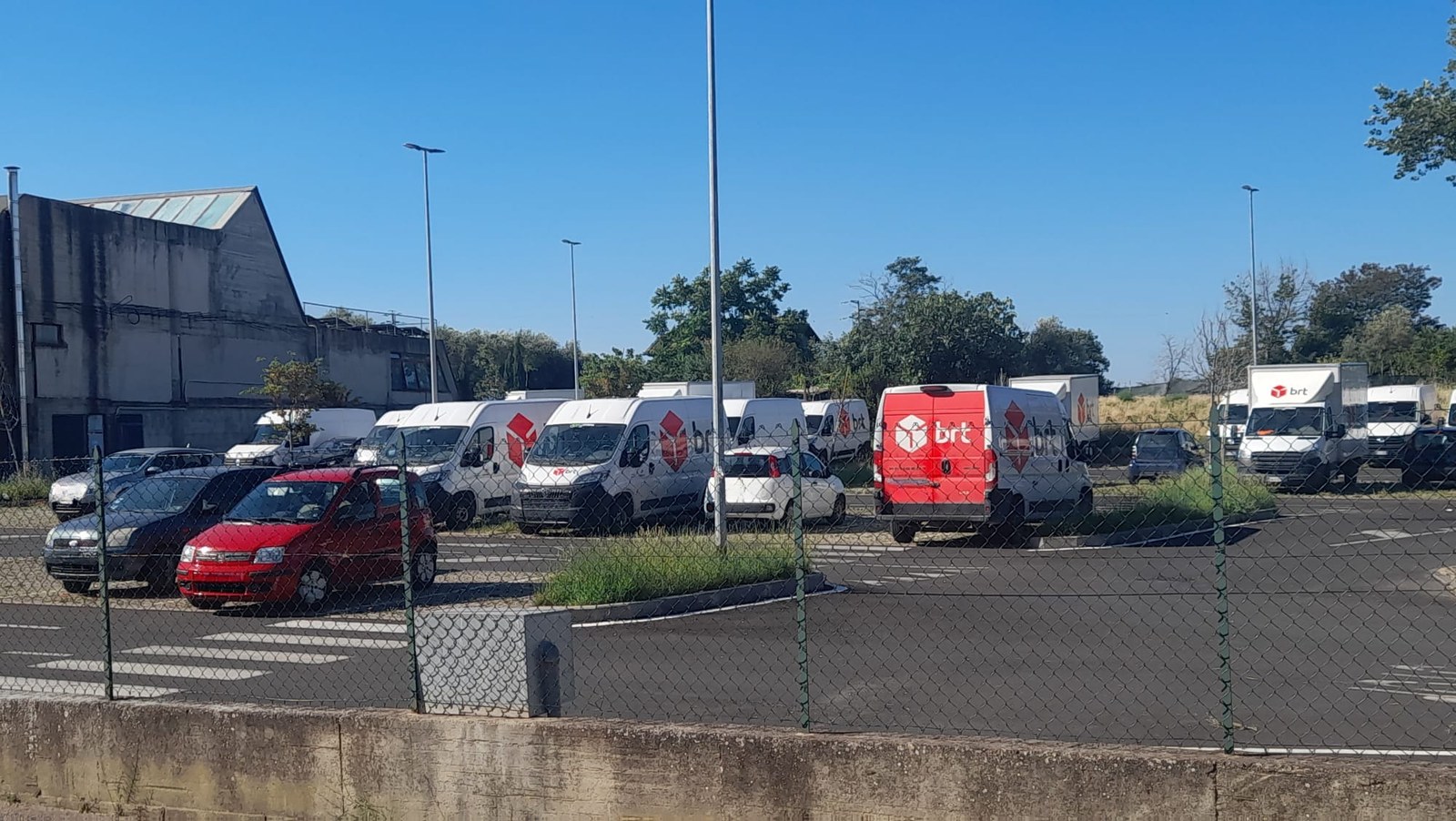Fila di furgoni bianchi e rossi con il marchio di un corriere espresso parcheggiati nel piazzale pubblico del Carmine a Viterbo, visti attraverso una recinzione
