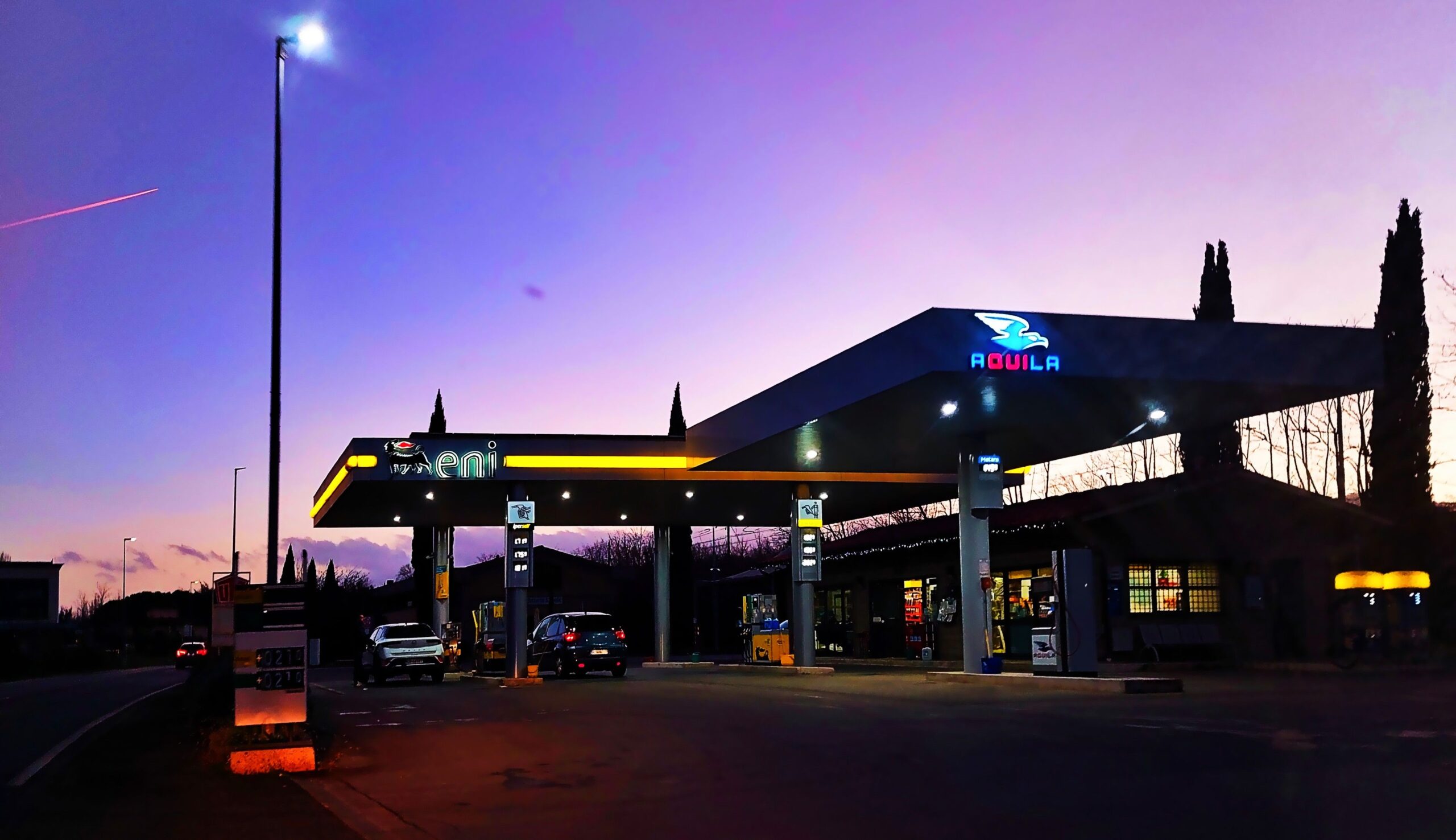 Distributore di carburante Eni illuminato al tramonto, con auto in sosta alle pompe