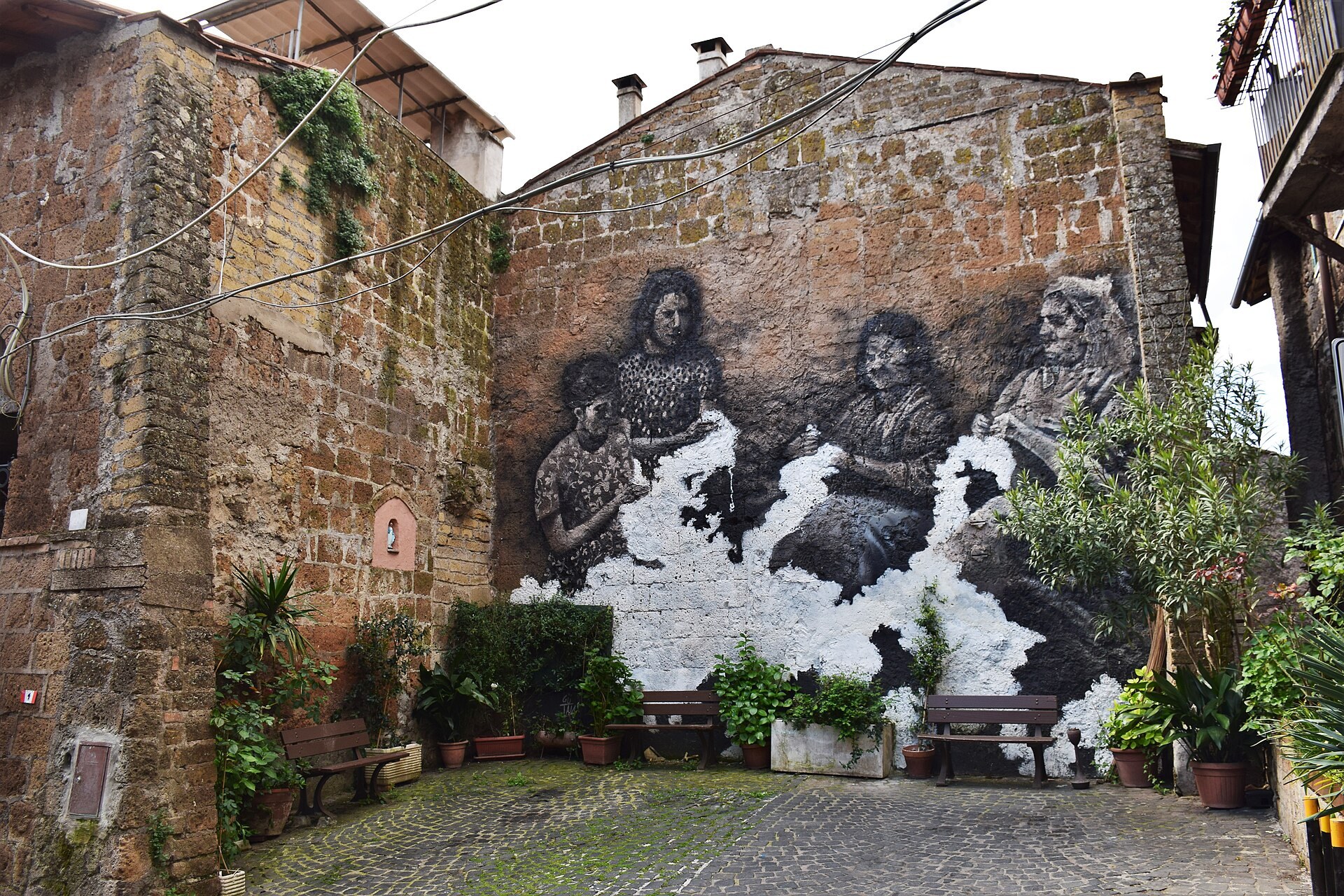 Un vicolo del borgo antico di Capranica con un grande murale su una parete di tufo, panchine e piante in vaso