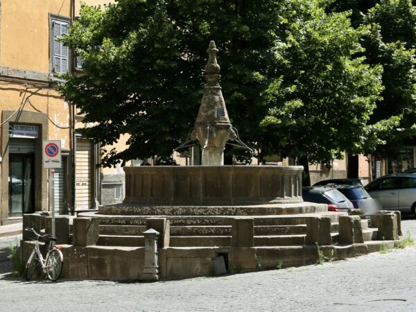 La fontana a fuso di San Faustino a Viterbo: vasca in peperino su gradini concentrici, fuso con teste di leone, piramide e pigna, alberi sullo sfondo