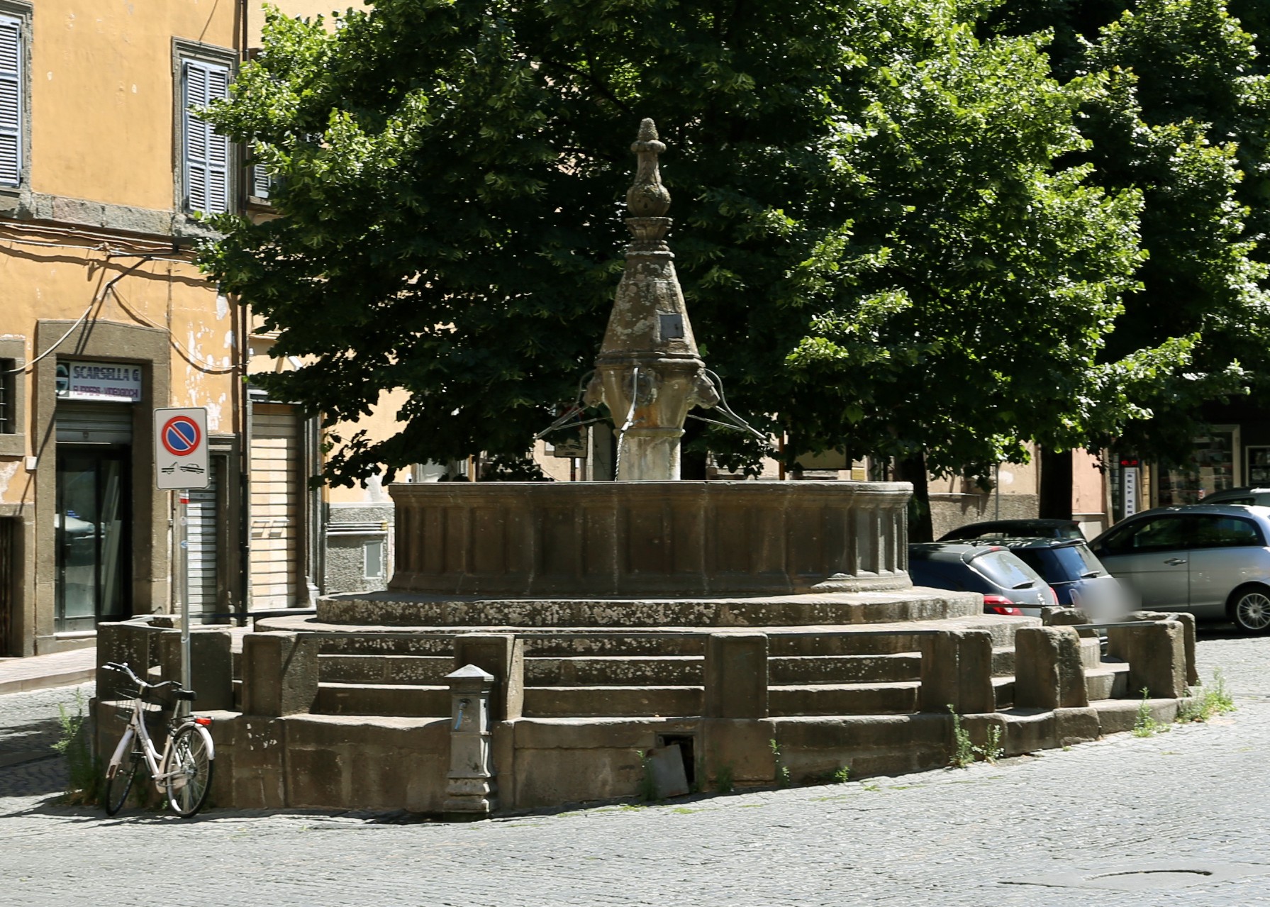 La fontana a fuso di San Faustino a Viterbo: vasca in peperino su gradini concentrici, fuso con teste di leone, piramide e pigna, alberi sullo sfondo