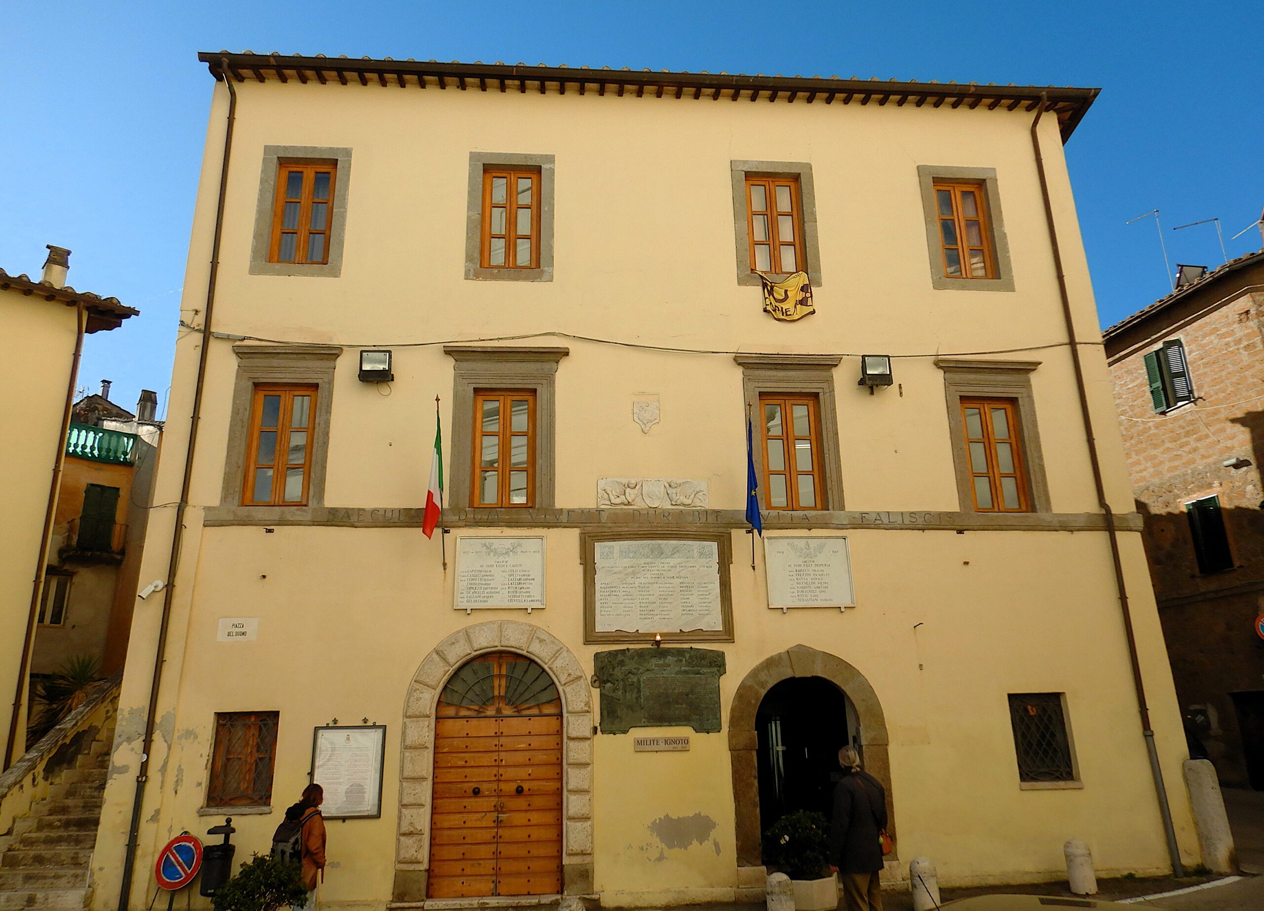 Facciata color ocra del Municipio di Gallese, con stemma comunale, bandiere italiana ed europea e portone d'ingresso ad arco