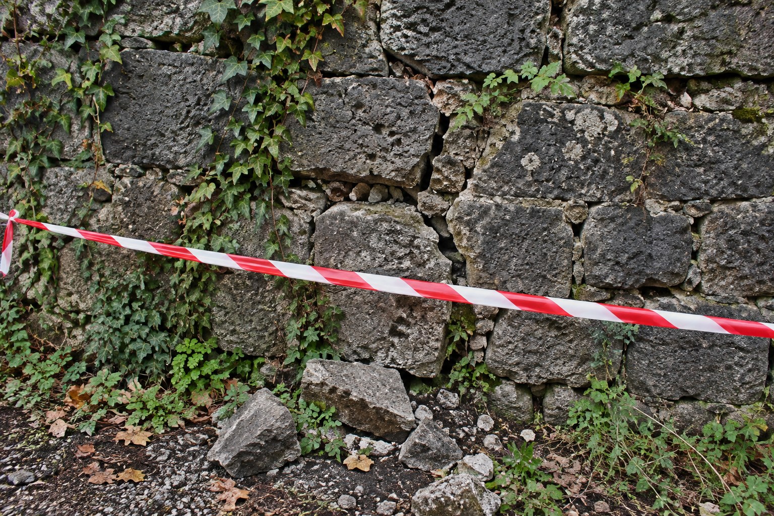 Muro in pietra grigia con crepe, edera e pietre cadute alla base, delimitato da un nastro bianco e rosso (immagine di repertorio)