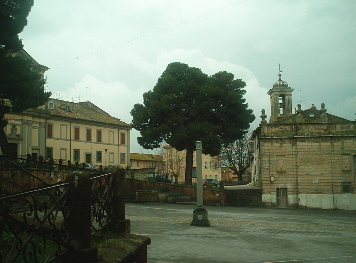 Piazza Dante a Monte Romano, con la chiesa, il grande pino al centro e i palazzi del paese sotto un cielo nuvoloso