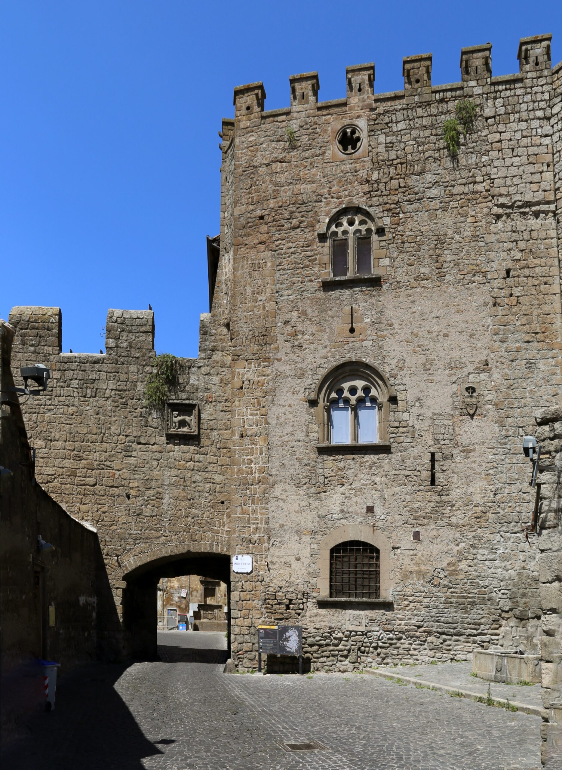 Porta San Pietro a Viterbo vista dall’interno: arco in peperino sotto la merlatura, con il leone scolpito in una cornice di pietra, accanto a un alto edificio merlato con bifore gotiche