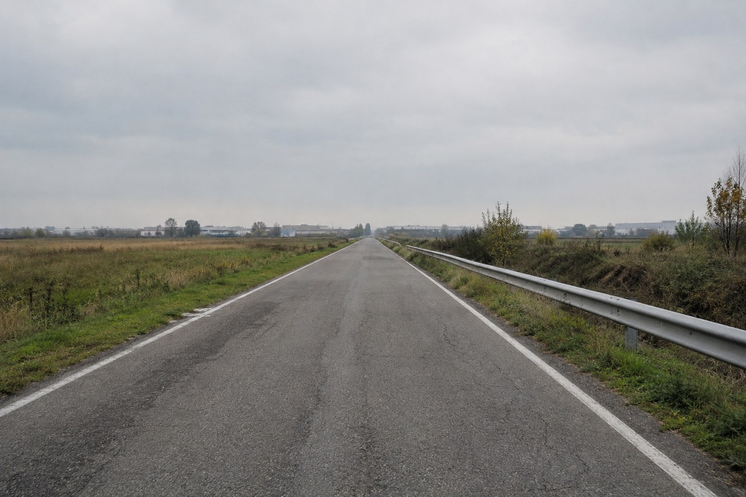 Strada asfaltata a due corsie tra campi incolti, con guardrail, in una giornata nuvolosa