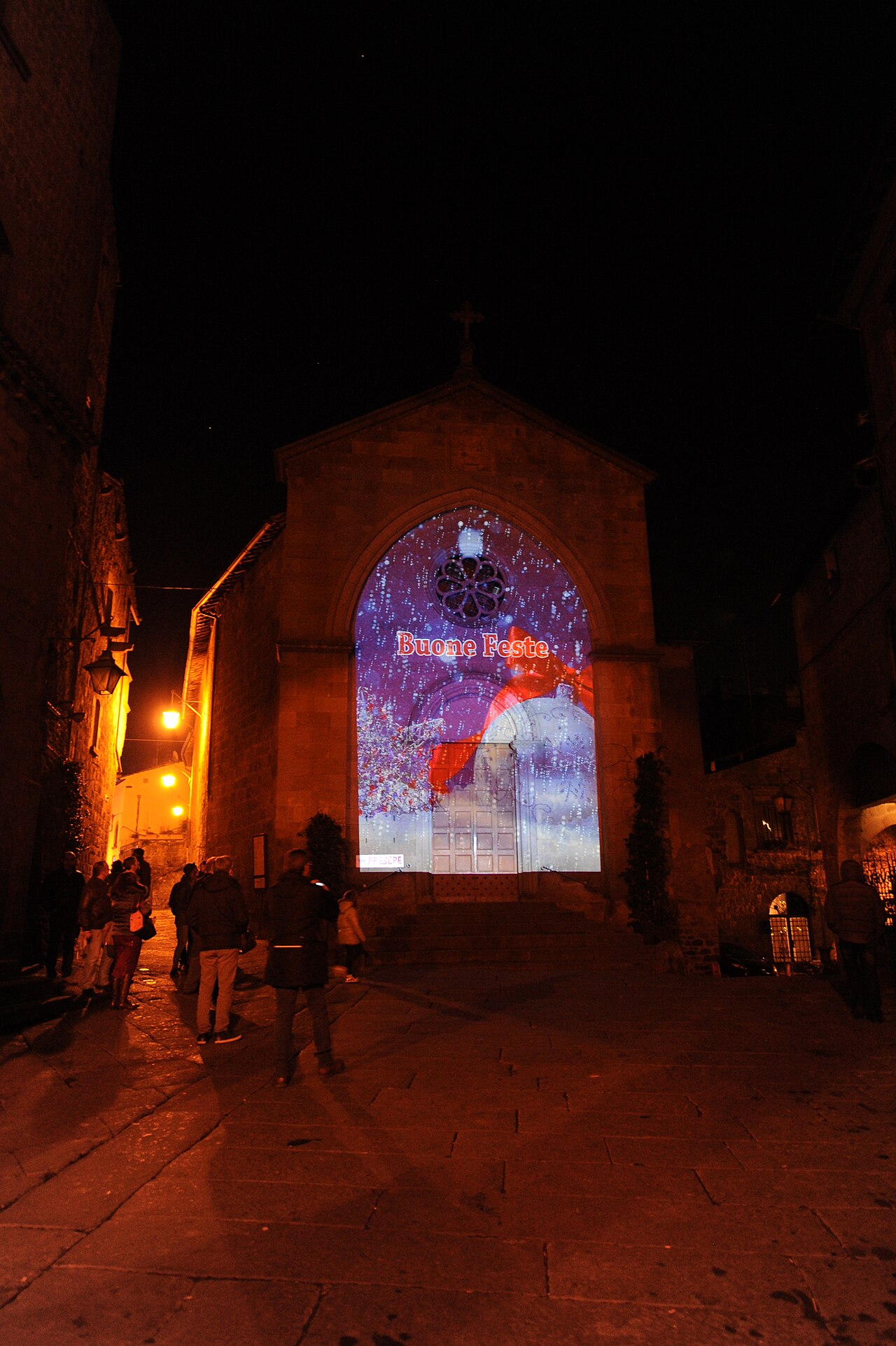 La facciata della chiesa di San Pellegrino illuminata di notte da una proiezione con la scritta Buone Feste, con alcuni passanti nella piazza